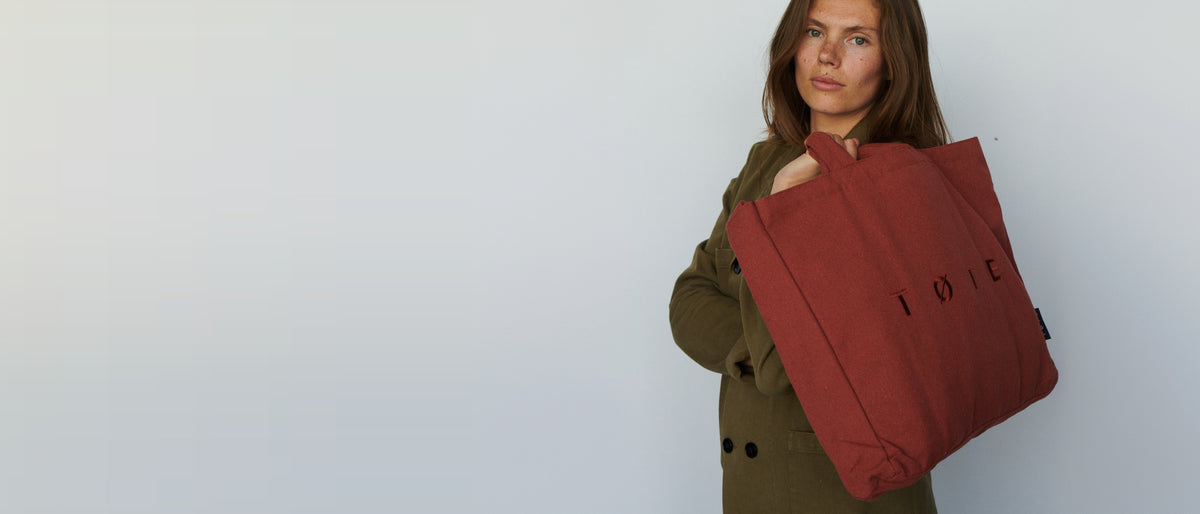 Model carrying a rust TOTE Bag against a light studio wall